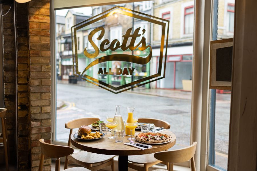 Window sign at Scott's All Day with a table laid for brunch behind, Cambridge